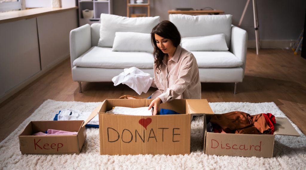 Woman sitting on living room floor sorting through old clothes into separate boxes to throw or donate